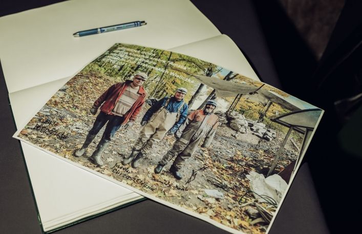 Leżąca fotografia przedstawiająca trzech mężczyzn w roboczych ubraniach przed wejściem do kopalni srebra. Zdjęcie leży na teczce, obok znajduje sie długopis. Osoby na zdjęciu są podpisane.