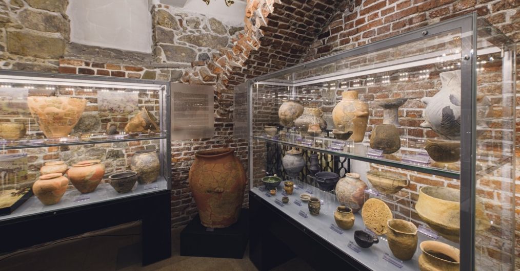 Fragment nowej wystawy archeologicznej w Zamku Żupnym. Widok na dwie, długie gabloty z naczyniami. Między nimi na podeście znajduje się duże naczynie zasobowe.