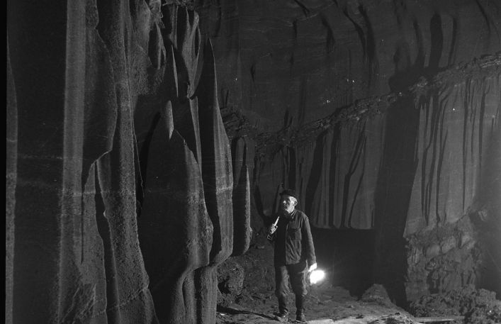 Czarno-biała fotografia przedstawiająca wnętrze kopalni soli. Obok solnej sciany stoi mężczyzna w roboczym ubraniu. Trzyma w ręce zapaloną lampę górniczą.