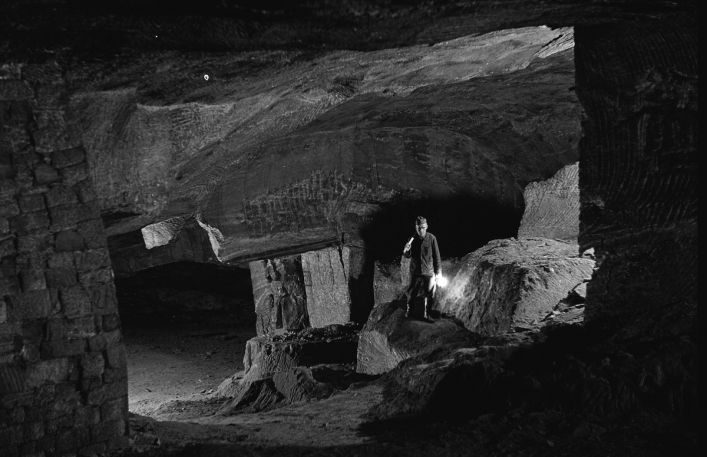 Czarno- biała fotografia pokazująca wnętrza kopalni. W oddali na tle solnych scian stoi człowiek z zapaloną lampą w ręce.