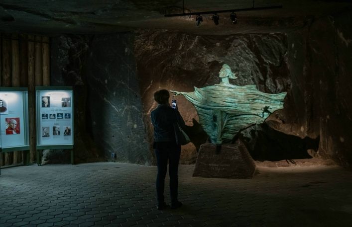 Turystka robi zdjęcie telefonem stojąc przed pomnikiem Fryderyka Chopina w kopalni soli. W tle wystawa pokazująca znane osoby zwiedzające kopalnię.