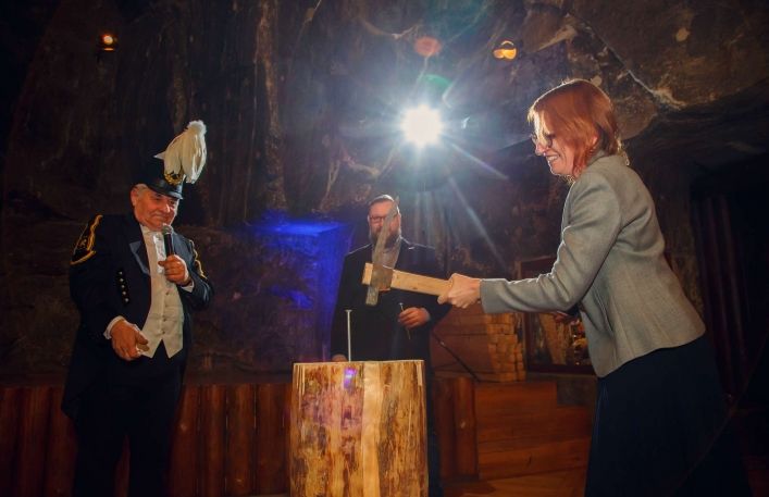 Kobieta próbuje wbić kilofem wielki gwóżdż do drewna. Obok górnik z mikrofonem i pracownik Muzeum Szymon Pawlikowski z drugim gwoździem w ręce.