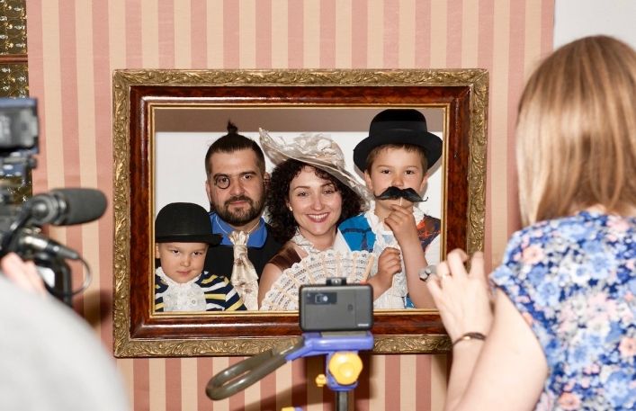 Wykonywanie pamiątkowej fotografii rodzinie w przebraniach. Czteroosobowa rodzina w kapeluszach, z wąsami oraz z monoklem pozuje w starej ramie obrazu.