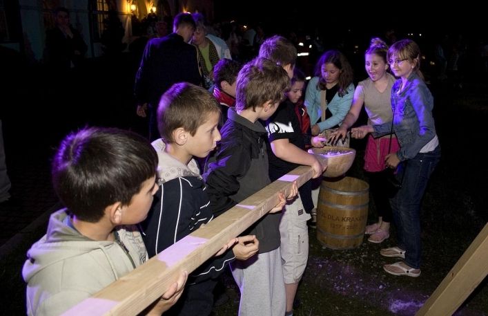Grupa dzieci trzyma w rękach ogromną, drewnianą łyżkę i wspólnie próbują przenieść na niej sól.