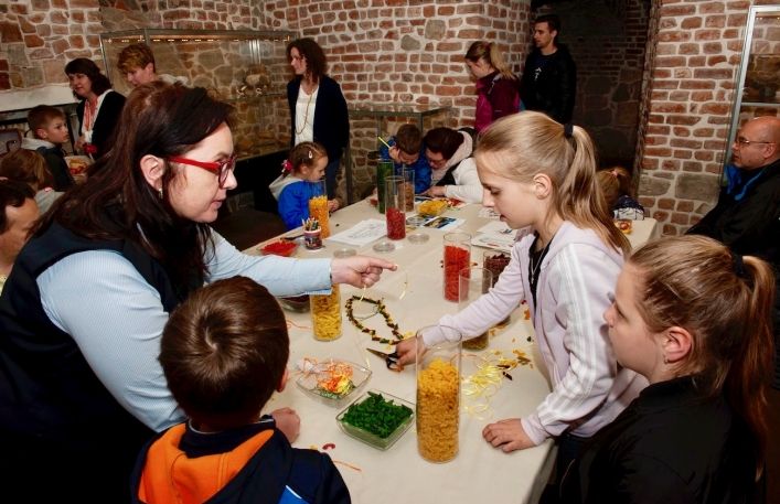 Zajęcia edukacyjne w Zamku. Edukatorka przy stole pokazuje dzieciom jak nawlekać kolorowe makarony na żyłkę.