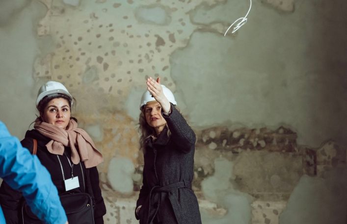 Klementyna Ochnika Dudek wskazuje ręką na polichromie i tłu,maczy coś stojącej obok dziennikarce.