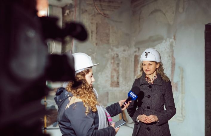 Dziennikaraka z TVP3 z mikrofonem w ręce, rozmawia z p. Klementyną Ochniak-Dudek- pracownicą Muzeum. W tle polichromia.