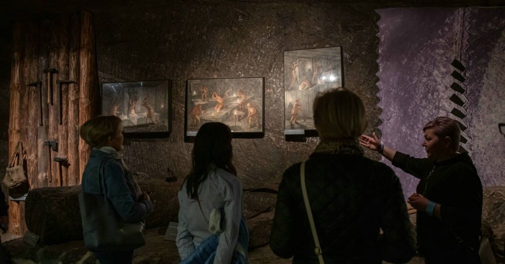 Przewodniczka objaśnia turystkom w jaki sposób tworzono bałwany solne na podstawie obrazów wiszących na ekspozycji.