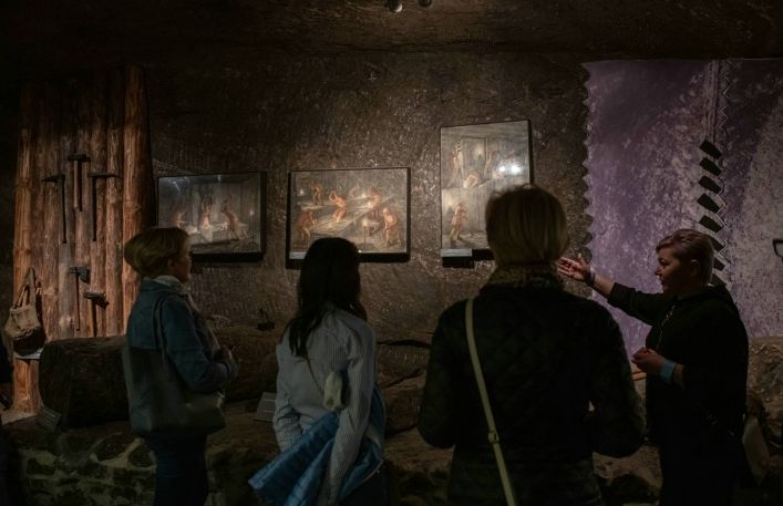 Przewodniczka objaśnia turystkom w jaki sposób tworzono bałwany solne na podstawie obrazów wiszących na ekspozycji.