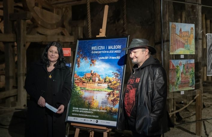 Uśmiechnięta autorka wystawy Magdalena Waśniowska Nowak stoi przy plakacie reklamującym wystawę. Obok jeden z artystów.