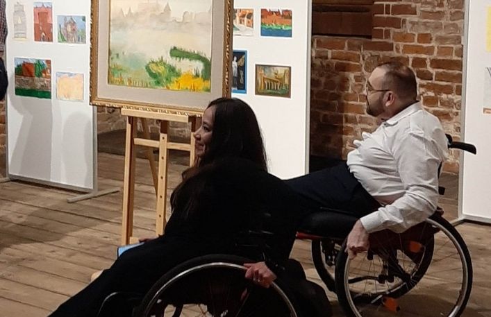 Dwie osoby- kobieta i mężczyzna na wózkach inwalidzkich zwiedzają wystawę.