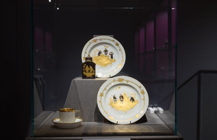 Gablota na wystawie. dwa okrągłe talerze ze złotymi dekoracjami i scenkami górniczymi w środku, herbatnica i filiżanka ze spodkiem.