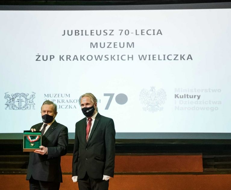 Minister Kultury Piotr Gliński  trzyma w ręce medal Gloria Artis obok niego stoi Dyrektor Muzeum na zdjęciu nie widać wyrazu twarzy, gdyż oboje są w maseczkach