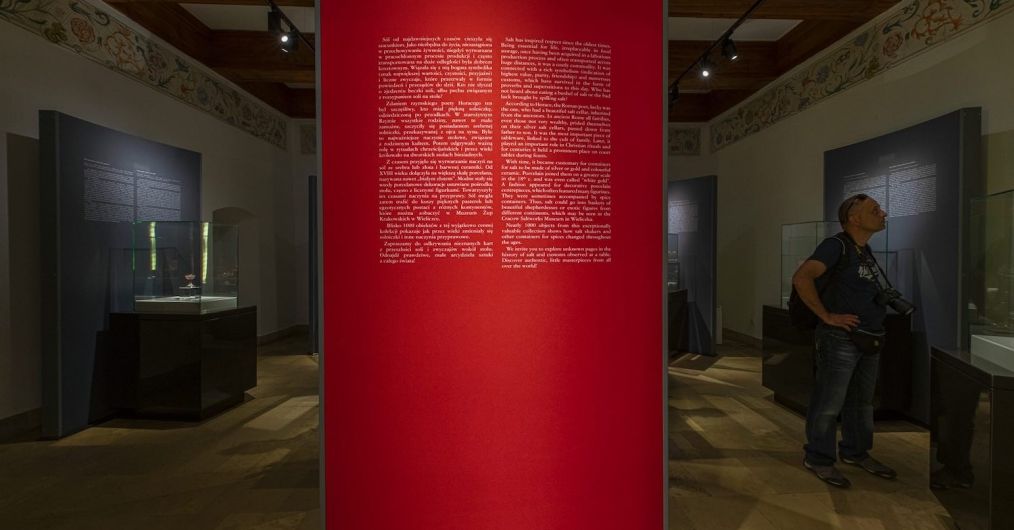 Widok na czerwona plansze z tekstem w wejściu na wystawę najcenniejszych solniczek w Zamku Żupnym. W tyle zwiedzający turysta.