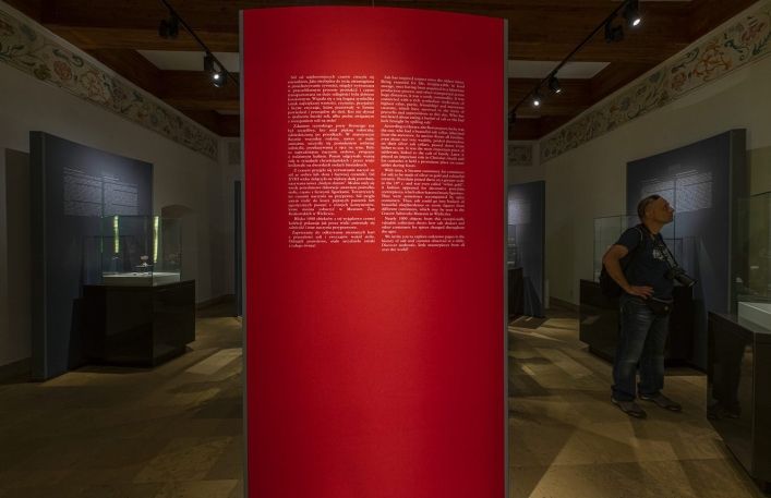 Widok na czerwona plansze z tekstem w wejściu na wystawę najcenniejszych solniczek w Zamku Żupnym. W tyle zwiedzający turysta.